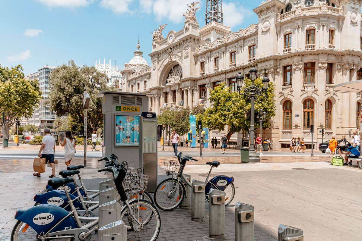 Fila di biciclette a noleggio Valenbisi presso una stazione di aggancio in una piazza di Valencia, con architettura storica e persone che camminano nelle vicinanze.
