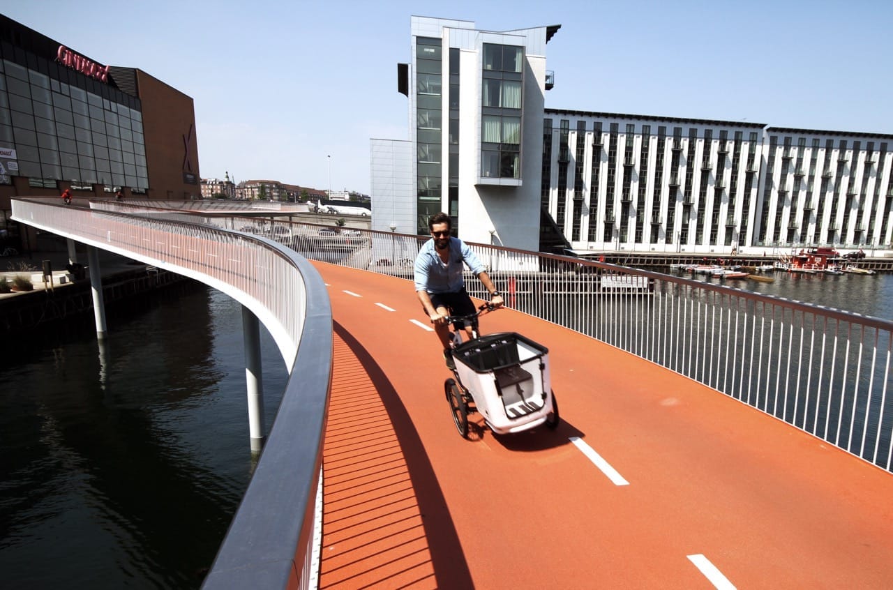 Il ponte ciclabile Cykelslangen a Copenaghen si curva sopra il porto, con un ciclista che percorre il suo percorso arancione brillante e moderni edifici sullo sfondo.