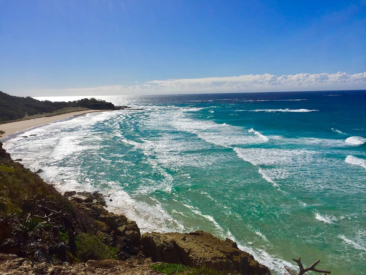 Blick auf den Cylinder Beach auf North Stradbroke Island mit türkisfarbenen Wellen, felsigen Klippen und einem breiten Sandstrand unter einem klaren blauen Himmel.