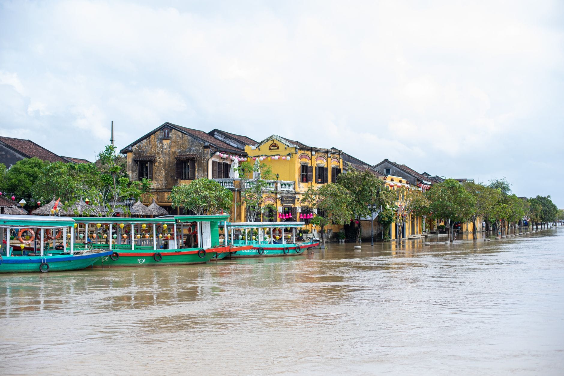 ทัศนียภาพริมแม่น้ำในเมืองฮอยอันพร้อมเรือหลากสีสัน อาคารประวัติศาสตร์สีเหลือง และต้นไม้เรียงรายตามแนวริมน้ำในเวลากลางวัน