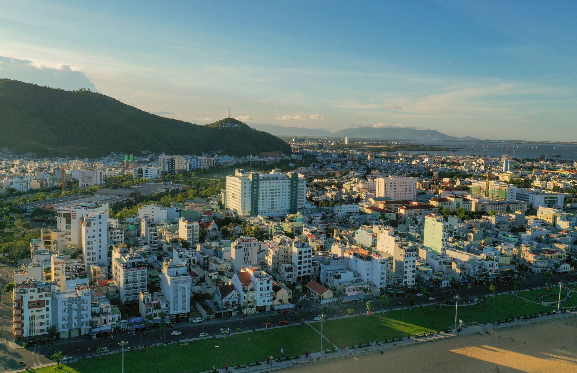 มุมมองทางอากาศของเมืองดานังพร้อมหาดทราย อาคารในเมือง และภูเขาที่อยู่ไกลออกไปภายใต้ท้องฟ้าสีฟ้าแจ่มใส