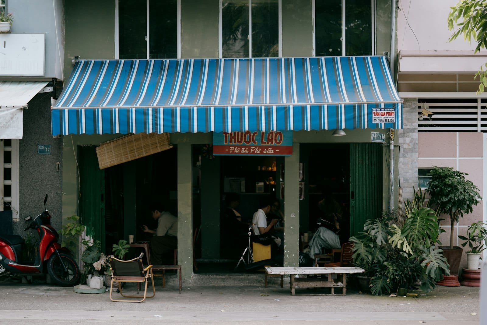 วิวถนนของร้านกาแฟเวียดนามที่มีกันสาดลายทางสีน้ำเงิน สกู๊ตเตอร์ และชาวบ้านที่นั่งอยู่ด้านนอกในดานัง