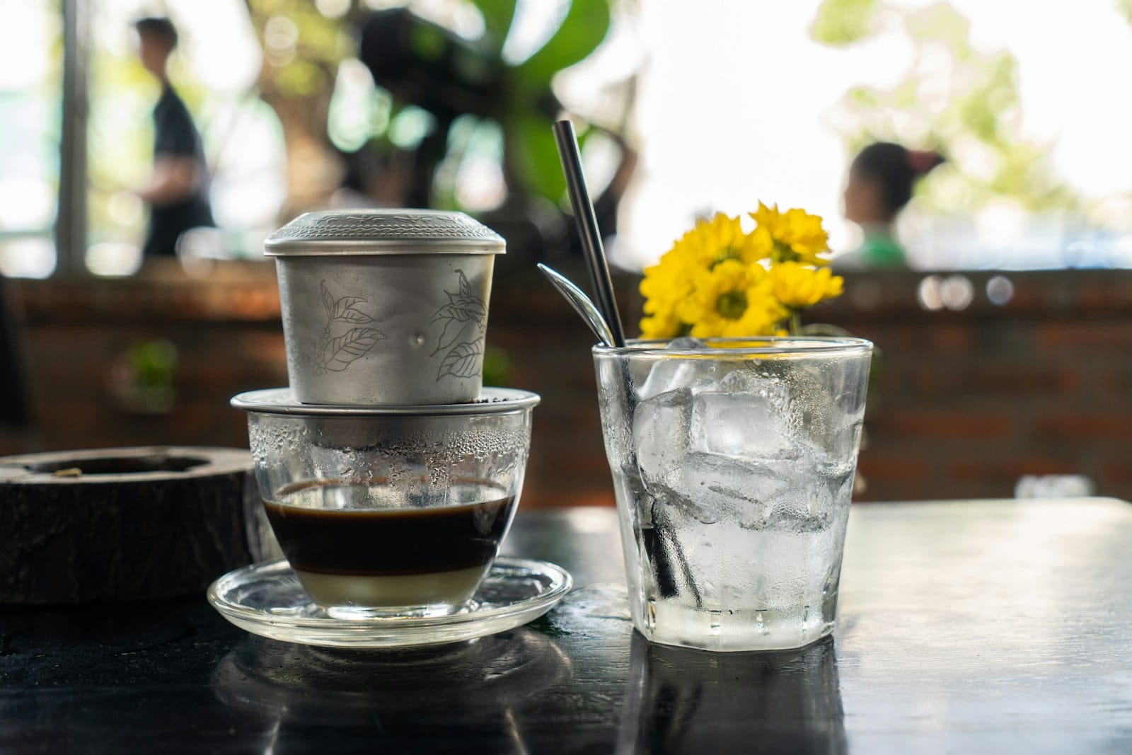 กาแฟเวียดนามที่กำลังดริปผ่านกรองฟินโลหะ วางข้างแก้วน้ำเย็นใส่น้ำแข็งและแจกันดอกไม้สีเหลืองใบเล็กบนโต๊ะคาเฟ่