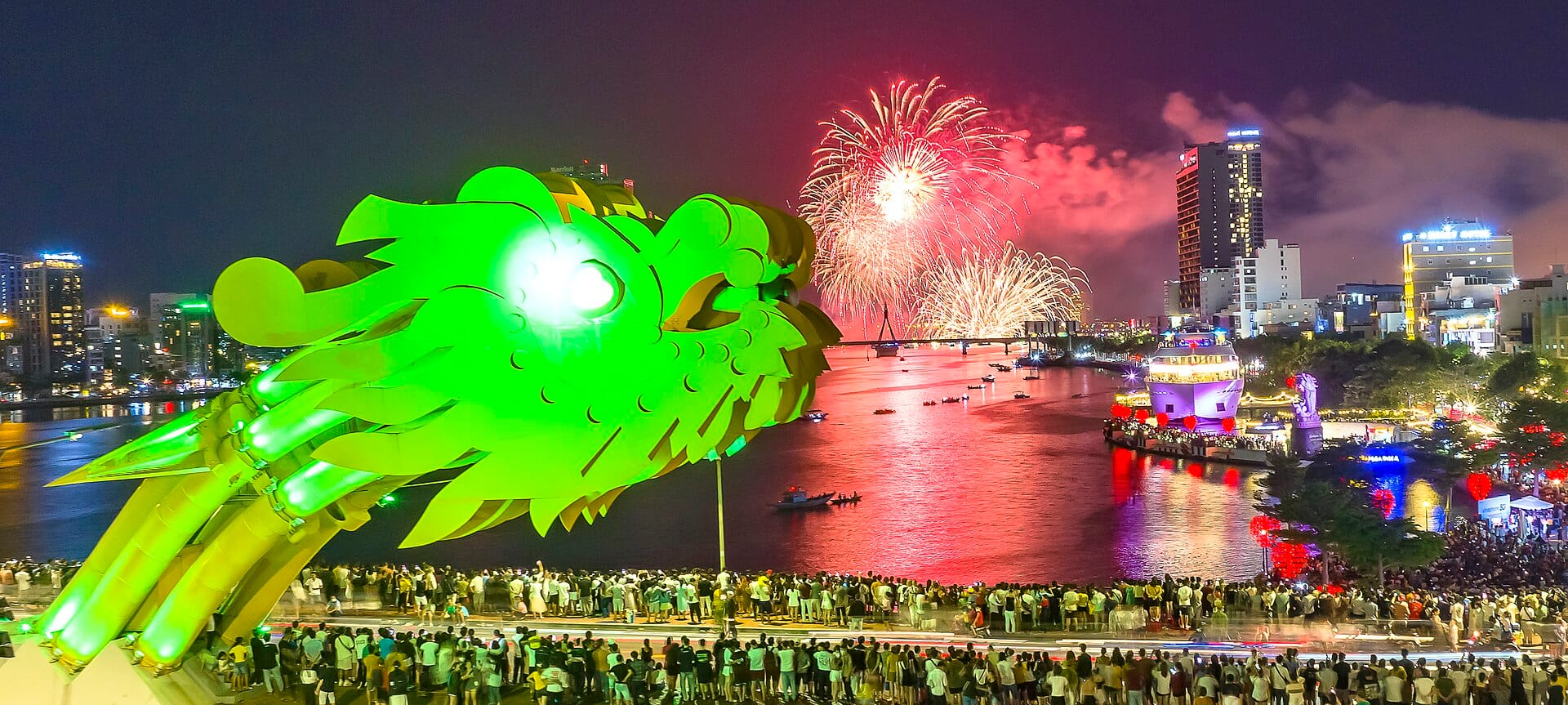 ฝูงชนจำนวนมากเรียงรายตามริมฝั่งแม่น้ำใต้ Dragon Bridge ของดานังในยามค่ำคืน ชมการแสดงดอกไม้ไฟอันตระการตาเหนือเส้นขอบฟ้าของเมืองและแม่น้ำฮัน