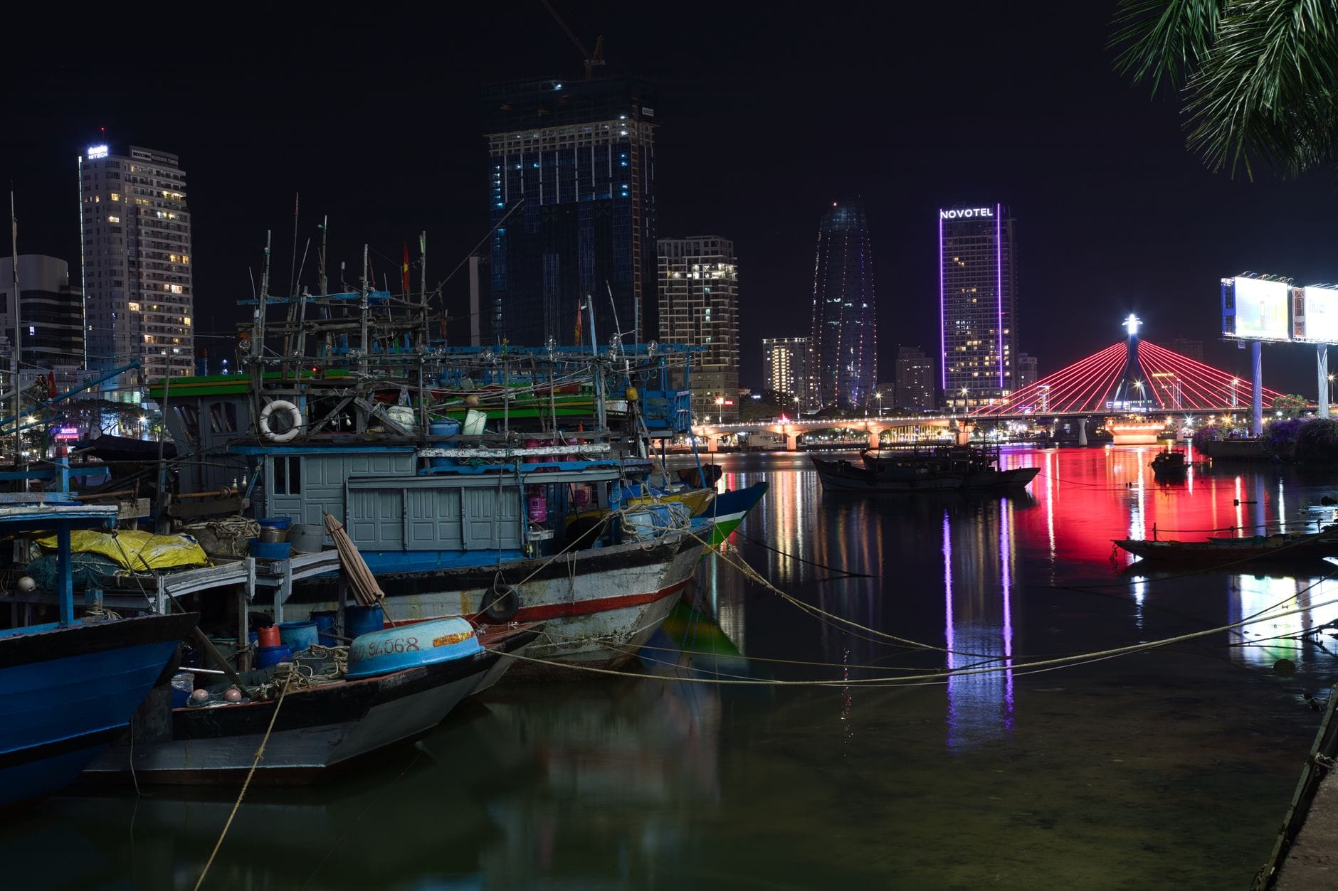 วิวกลางคืนของโรงแรมริมแม่น้ำและเส้นขอบฟ้าของดานังพร้อมไฟสีสันสดใสและเรือแบบดั้งเดิมที่จอดเทียบท่าอยู่เบื้องหน้า