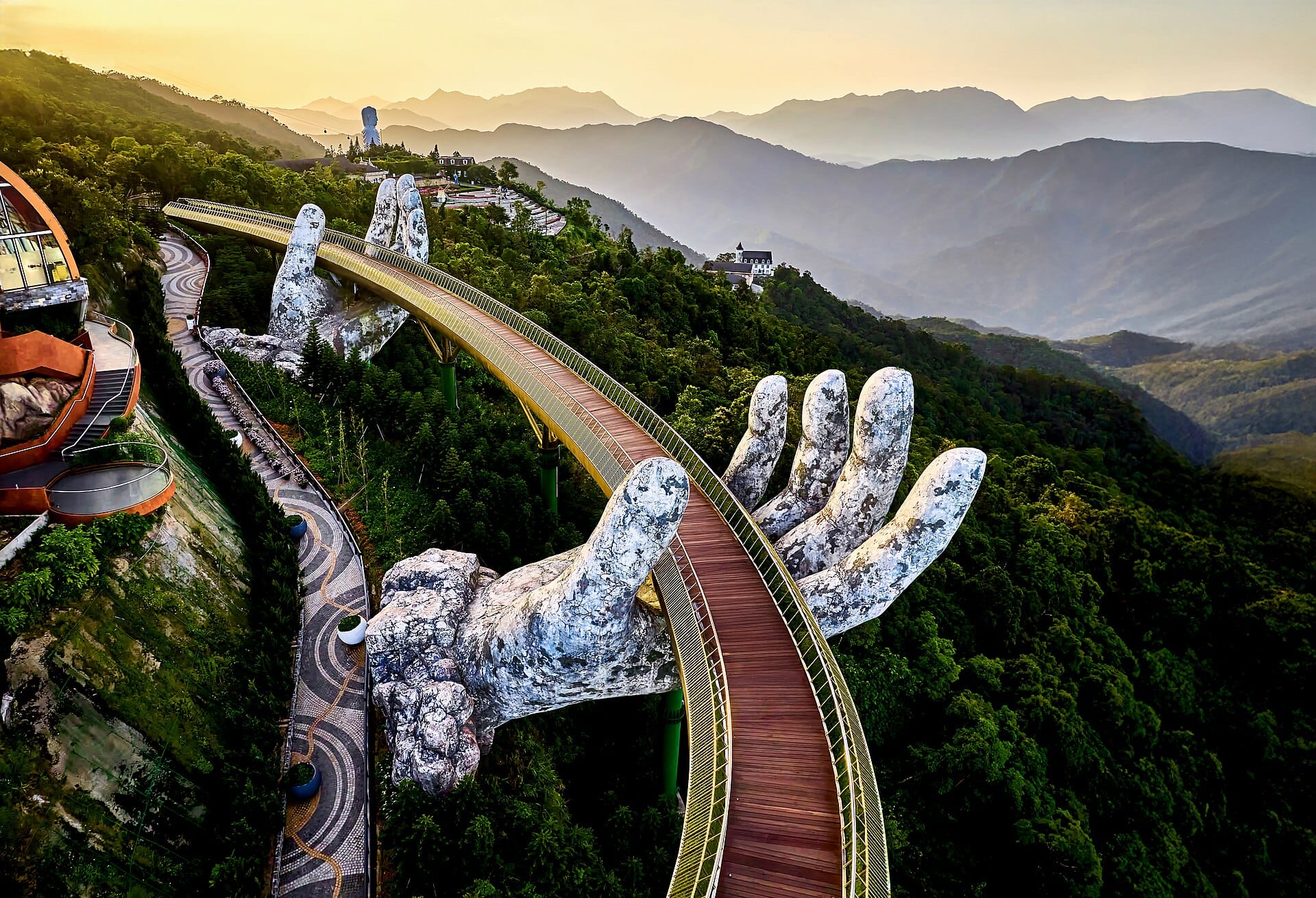 ภาพทางอากาศของ Golden Bridge ที่ Ba Na Hills ในดานัง โดดเด่นด้วยมือหินยักษ์รองรับทางเดินโค้งเหนือเทือกเขาสีเขียวขจีในยามพระอาทิตย์ตกดิน