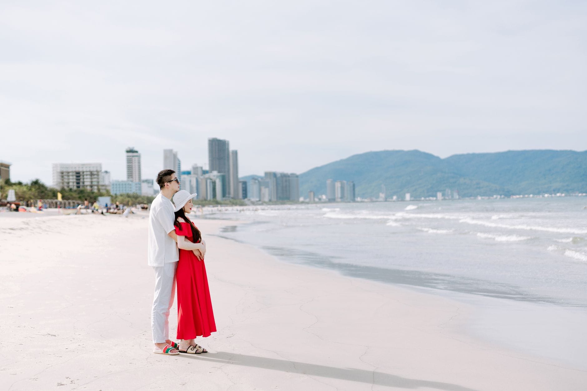 คู่รักยืนริมหาดดานัง มีเส้นขอบฟ้าเมืองและภูเขาเขียวด้านหลัง ใต้ท้องฟ้าสดใส