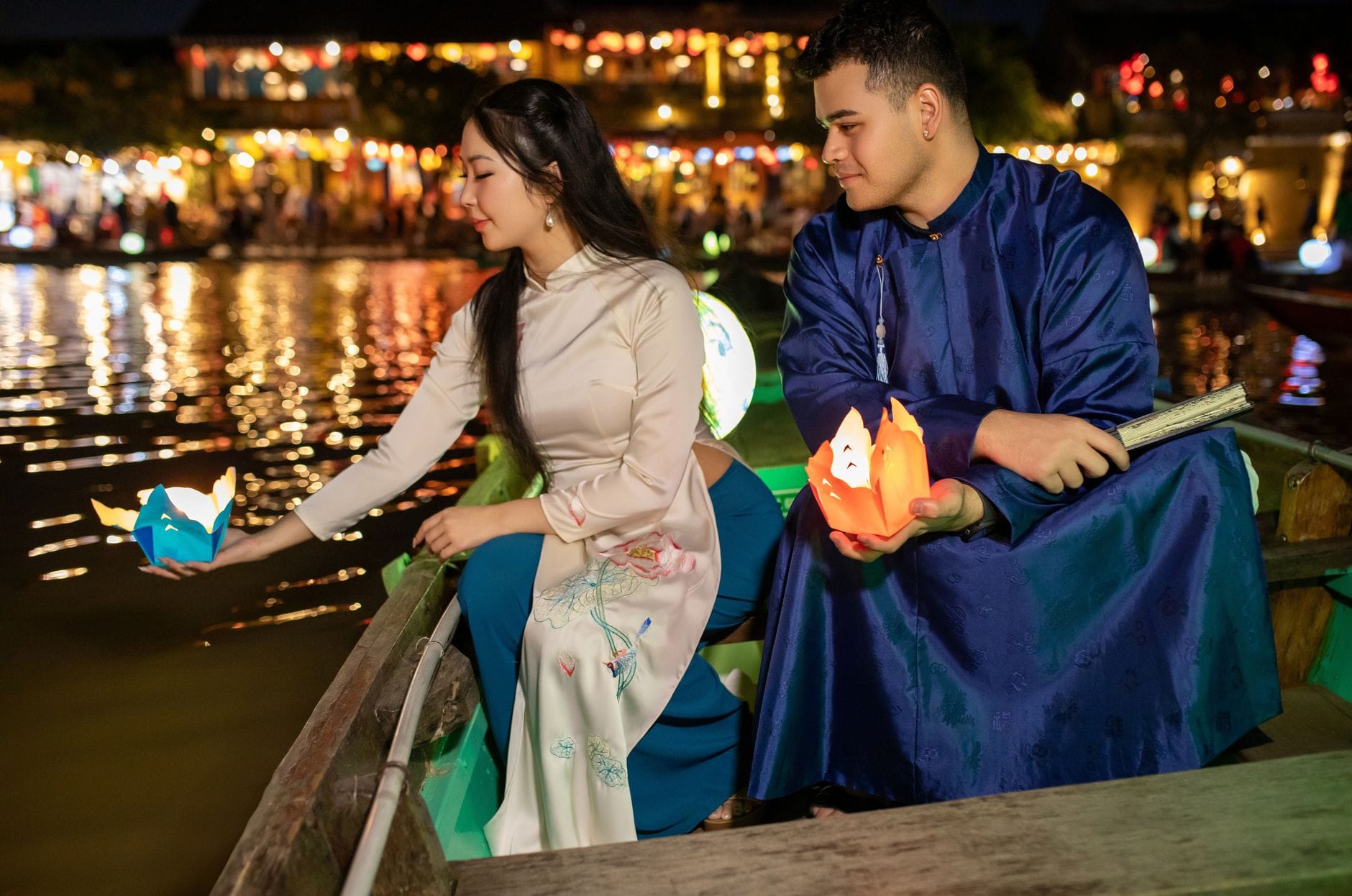 คู่รักสวมชุดพื้นเมืองนั่งอยู่ในเรือที่ประดับโคมไฟยามพลบค่ำบนแม่น้ำในฮอยอัน กำลังปล่อยโคมเทียนลงสู่ผิวน้ำ