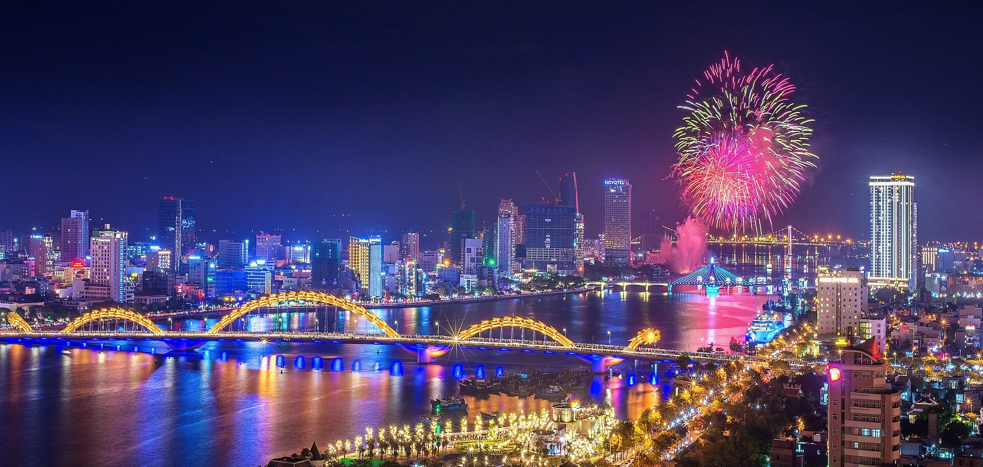 วิวกลางคืนอันสดใสของแม่น้ำฮานในดานัง พร้อมด้วย Dragon Bridge ที่สว่างไสว เส้นขอบฟ้าของเมือง ดอกไม้ไฟ และเงาสะท้อนบนผิวน้ำ
