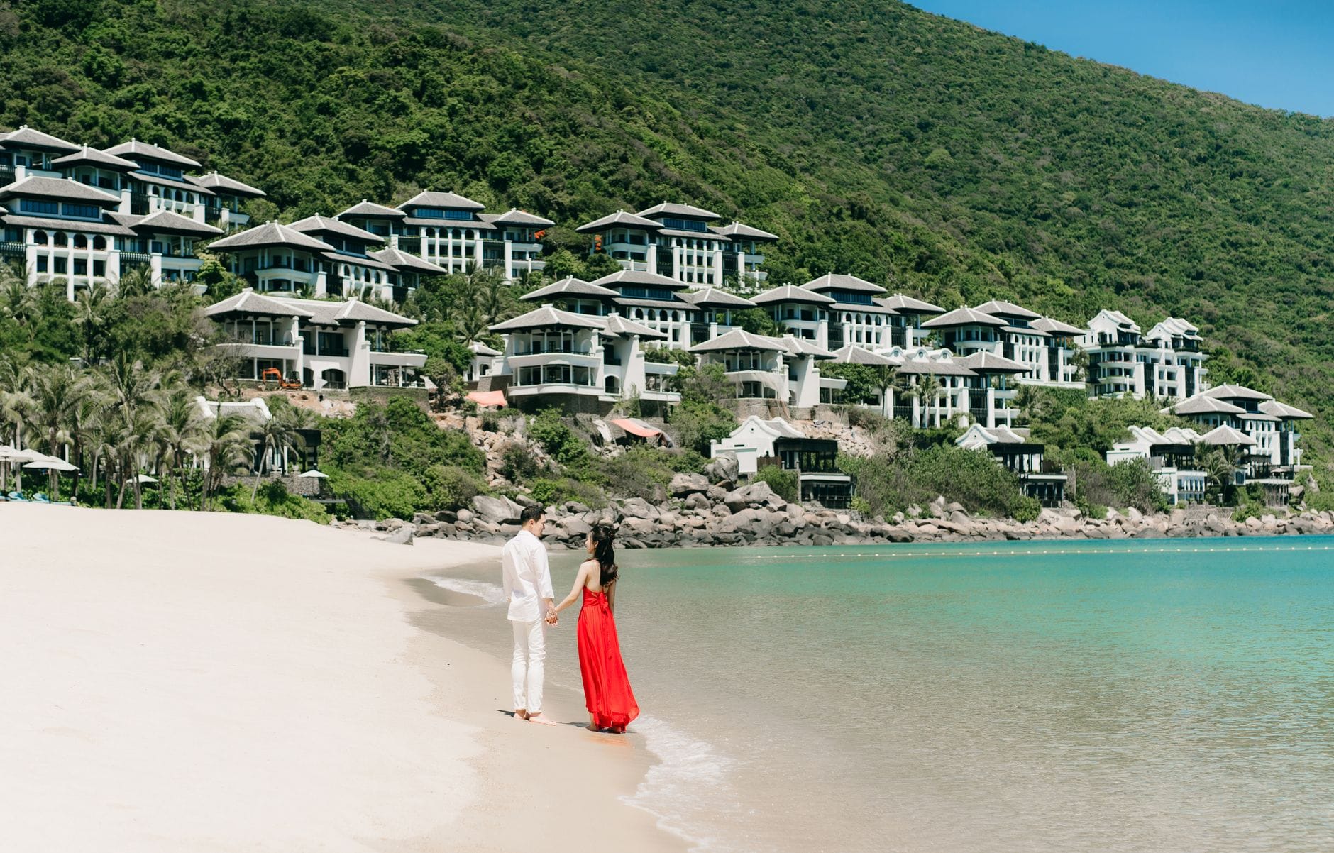 คู่รักบนชายหาดทรายขาวเบื้องหน้าอาคารรีสอร์ทหรูบนเนินเขาเหนือน้ำสีเทอร์คอยส์และเนินเขาสีเขียวชอุ่มใน Da Nang