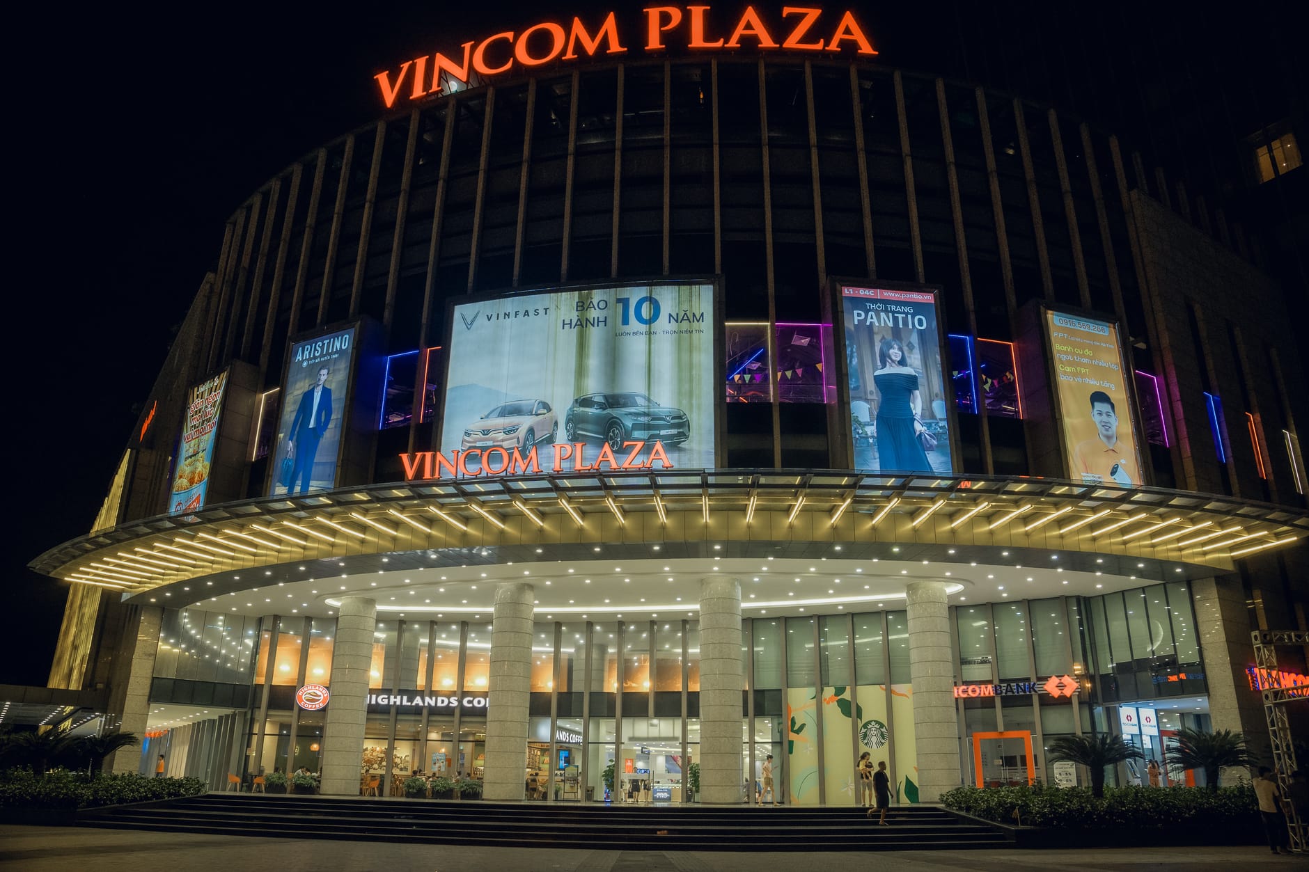 วิวยามค่ำคืนของทางเข้า Vincom Plaza Da Nang สว่างไสวพร้อมร้านค้าและโฆษณาแฟชั่นที่มองเห็นได้ชัดเจน