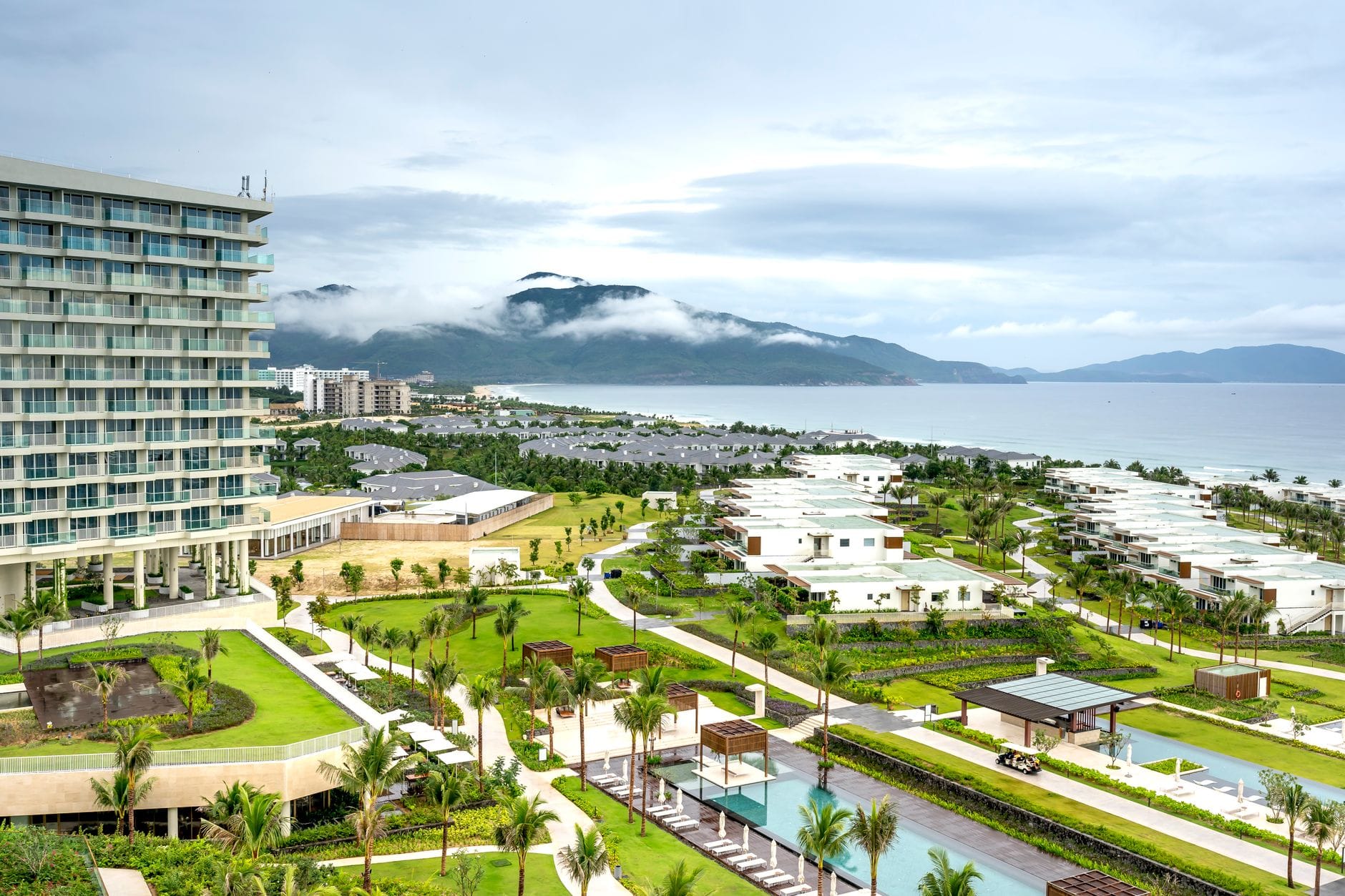 รีสอร์ทริมชายหาดและวิลล่าสมัยใหม่พร้อมสระว่ายน้ำตลอดแนวชายฝั่งของดานัง พร้อมฉากหลังเป็นภูเขาและวิวทะเลในวันที่มีเมฆครึ้ม