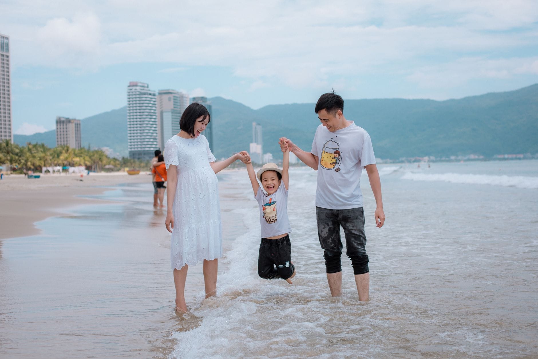 ครอบครัวกับลูกเล็กเล่นน้ำตื้นบนชายหาดทรายดานัง มีวิวเมืองและภูเขาสีเขียวอยู่ด้านหลัง