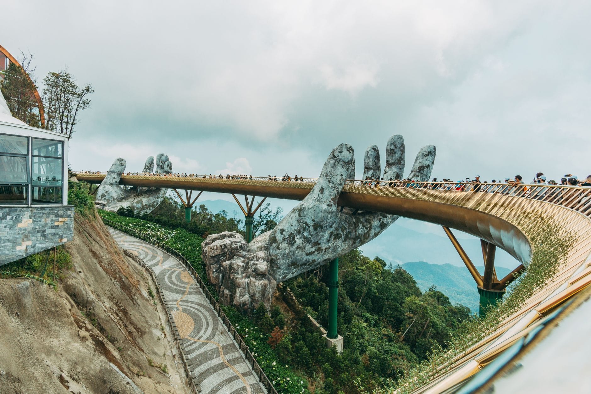 สะพาน Golden Bridge ใน Da Nang สะพานสำหรับคนเดินเท้าที่ถูกประคองโดยมือหินยักษ์สองข้าง ล้อมรอบด้วยภูเขาและพื้นที่สีเขียวใต้ท้องฟ้าที่มีเมฆมาก