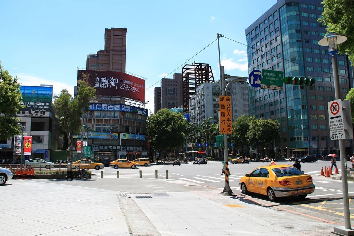 Sonniger Tag im Bezirk Da'an, Taipeh, mit modernen Bürogebäuden, grünen Bäumen, gelben Taxis und belebten Stadtstraßen.