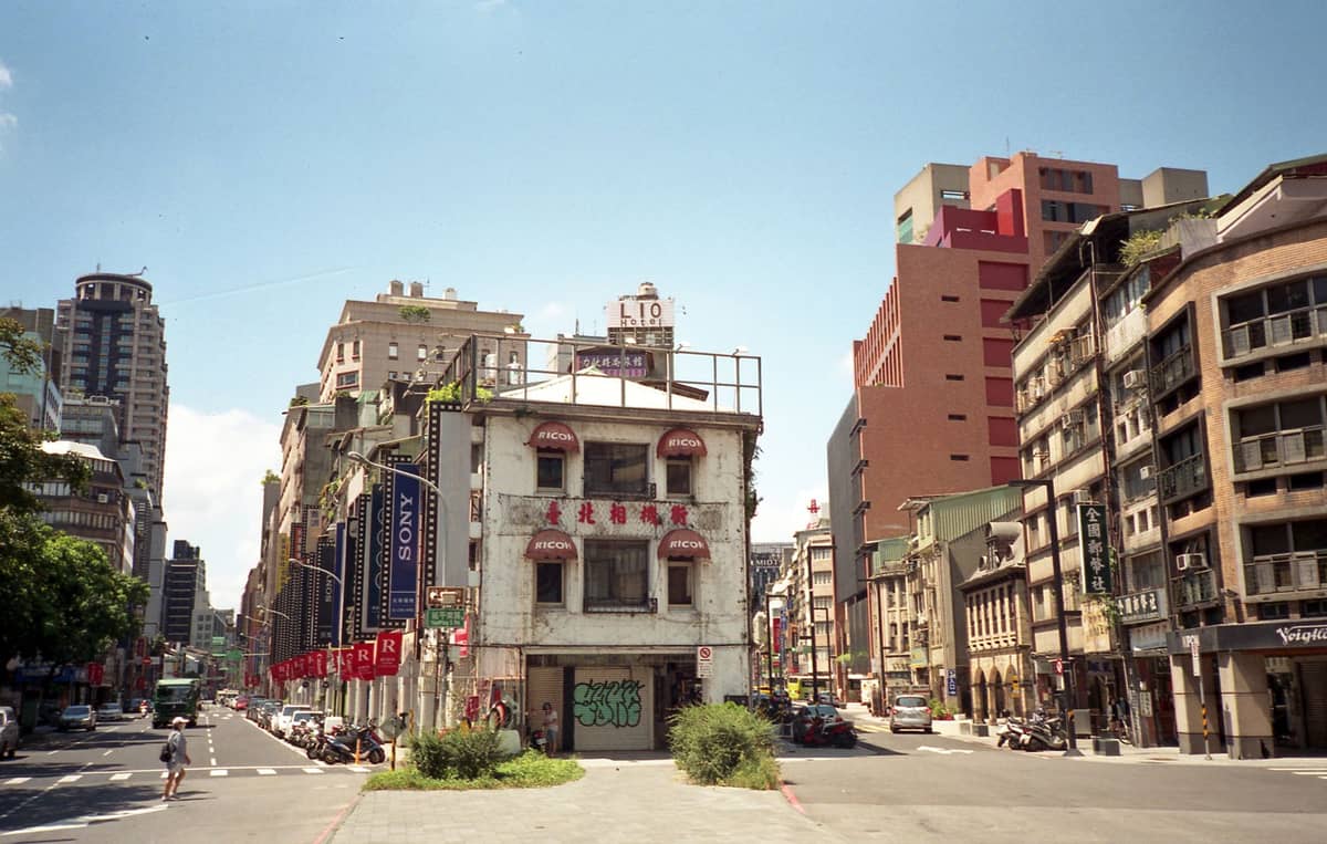 Kreuzung in Dadaocheng mit einem Ladenhaus im typischen Stil, umgeben von mittelhohen Gebäuden und Bannern unter einem klaren Mittagshimmel in Taipeh.