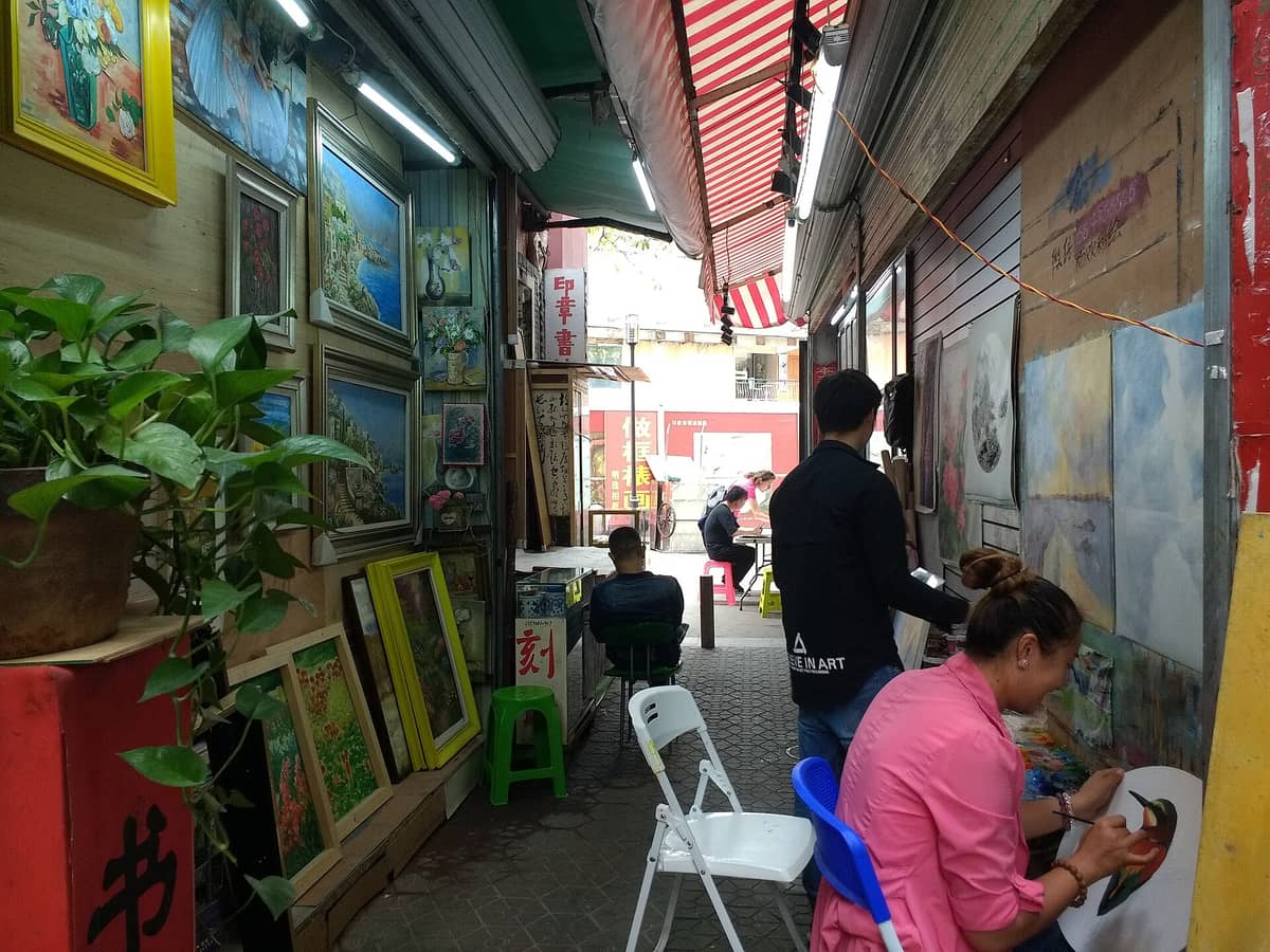 Um beco estreito na Vila de Pintura a Óleo de Dafen, com as paredes repletas de quadros coloridos emoldurados, artistas trabalhando e uma atmosfera animada e convidativa de ateliê de arte.