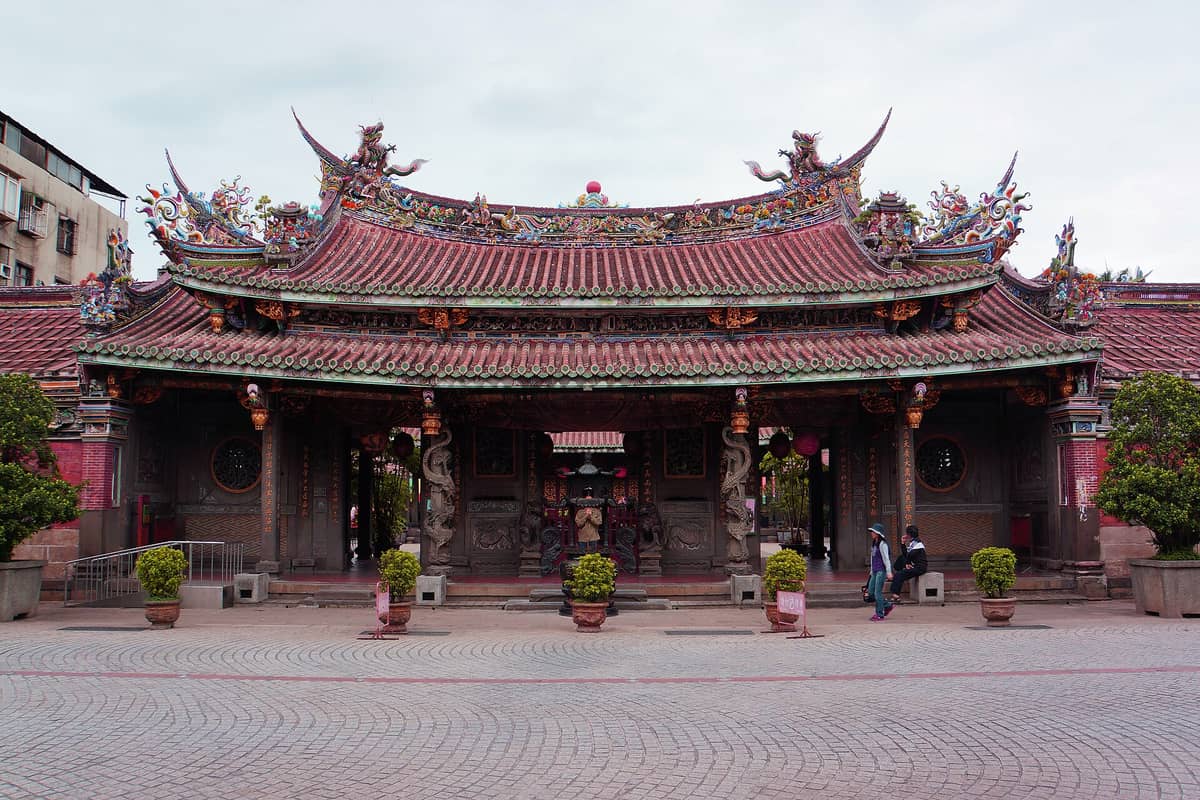 Frontansicht des Dalongdong Baoan-Tempels in Taipeh mit kunstvollen traditionellen chinesischen Dachverzierungen, roten Säulen und Topfpflanzen entlang des Tempeleingangs.