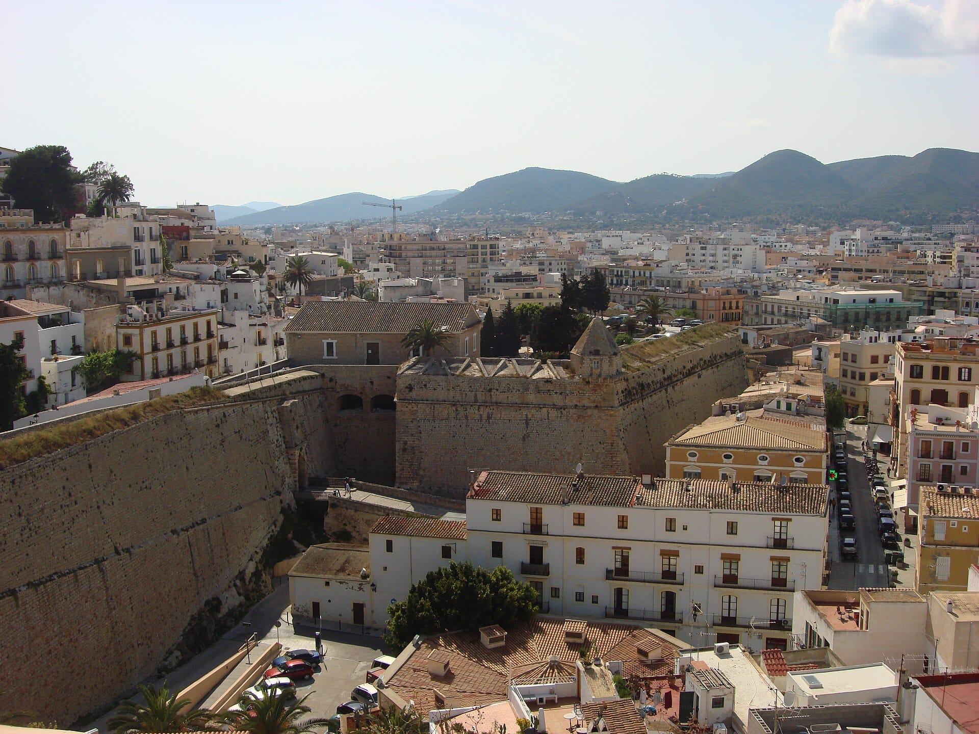 Vista delle imponenti mura in pietra della fortezza di Dalt Vila circondate da edifici storici a Ibiza Town, con le montagne sullo sfondo.