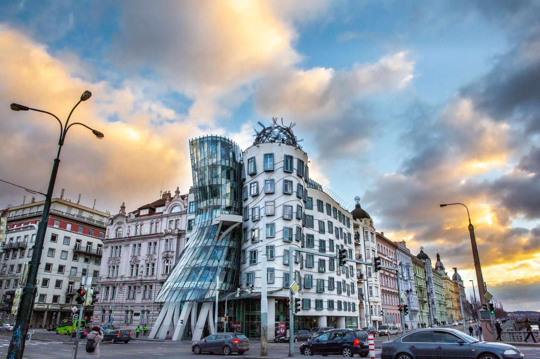Das Dancing House in Prag bei Sonnenuntergang fotografiert, mit dramatischen Wolken und warmem Abendlicht, das die einzigartige, geschwungene Architektur des Gebäudes auf einer belebten Stadtstraße hervorhebt.