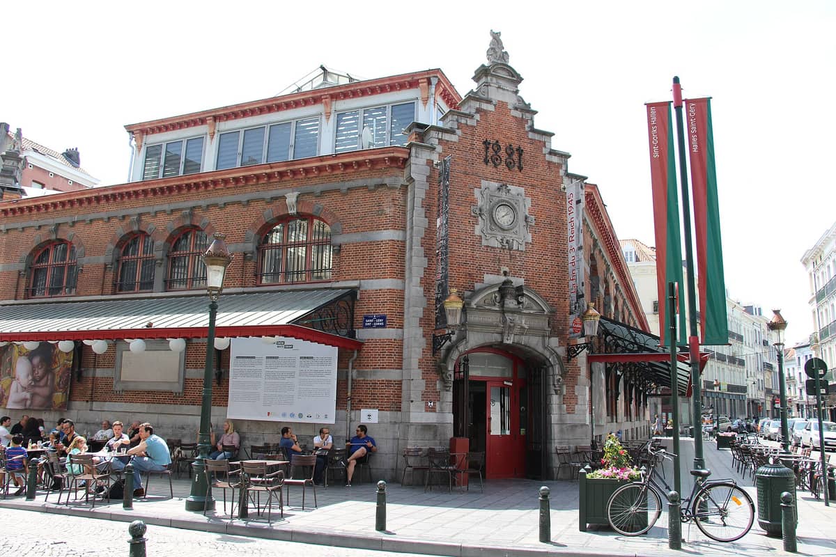 Fachada das históricas Halles Saint-Géry em Bruxelas, com tijolos vermelhos, janelas em arco, mesas de café ao ar livre e pessoas animando a praça.