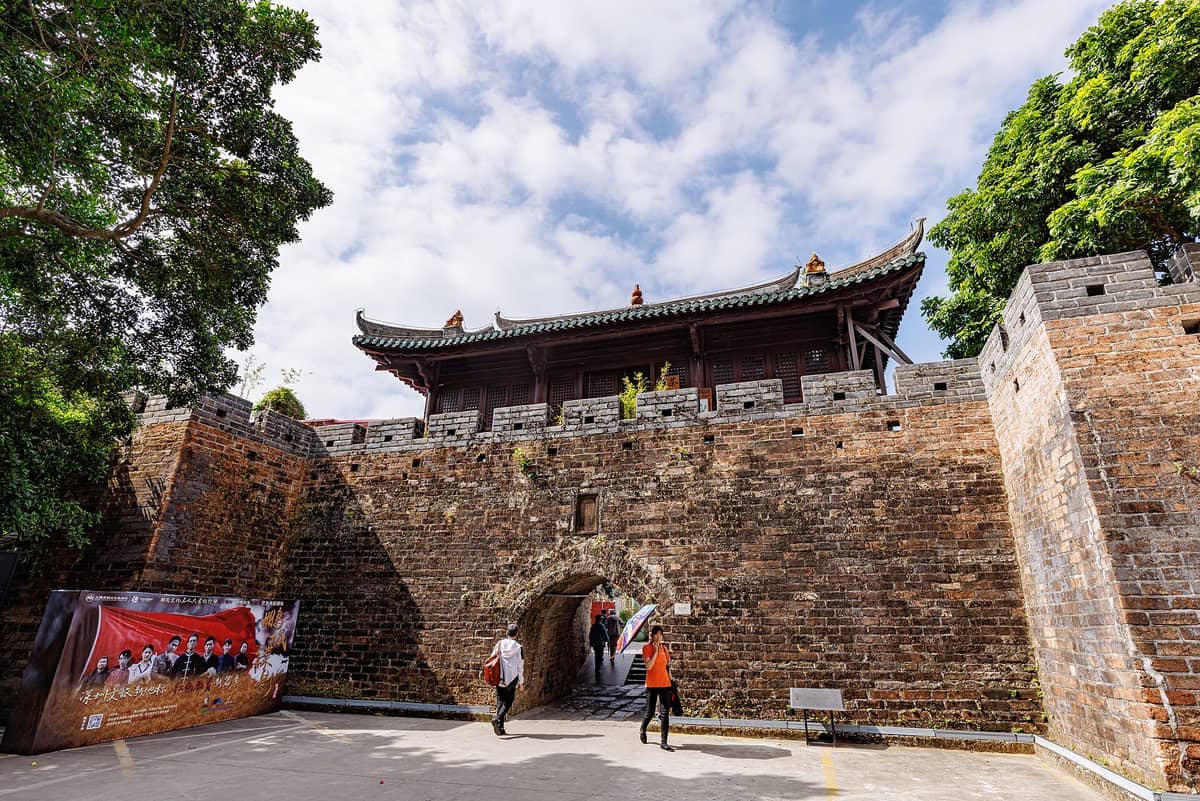 Entrada principal da Fortaleza Antiga de Dapeng com grossas paredes de pedra, telhado tradicional de telhas, árvores e visitantes passando sob um céu azul.
