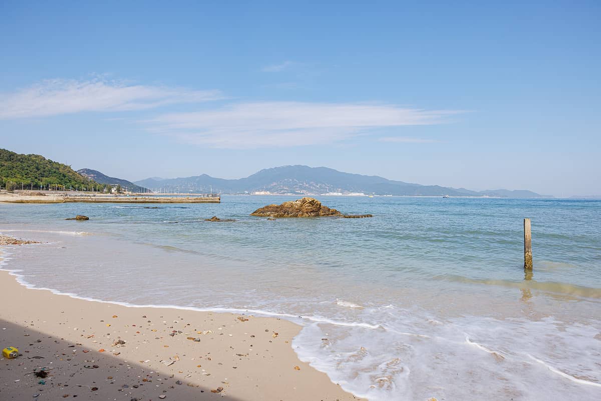 Uma praia de areia com rochas espalhadas, ondas suaves e vista para colinas arborizadas e montanhas ao longe na Península de Dapeng, em Shenzhen.