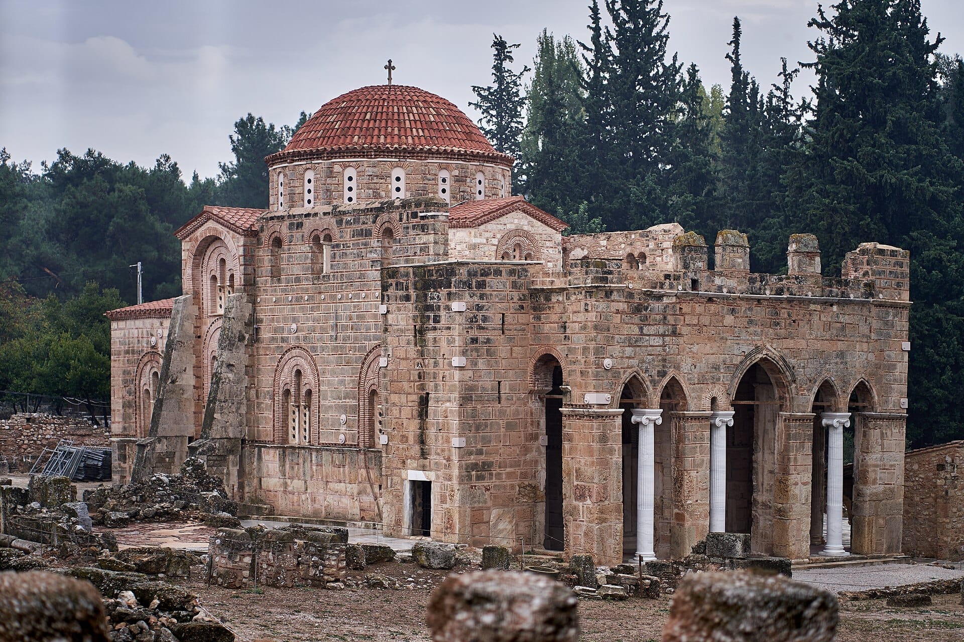 Il Monastero di Daphni con la sua distintiva cupola bizantina, archi in pietra e colonne, circondato da vegetazione e rovine antiche sotto un cielo nuvoloso.