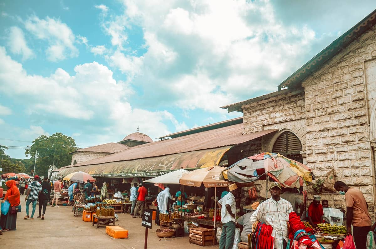 Scena vivace fuori dal Mercato Darajani a Stone Town: l'edificio storico, i venditori, gli ombrelloni colorati e gli acquirenti sotto un cielo azzurro con qualche nuvola.
