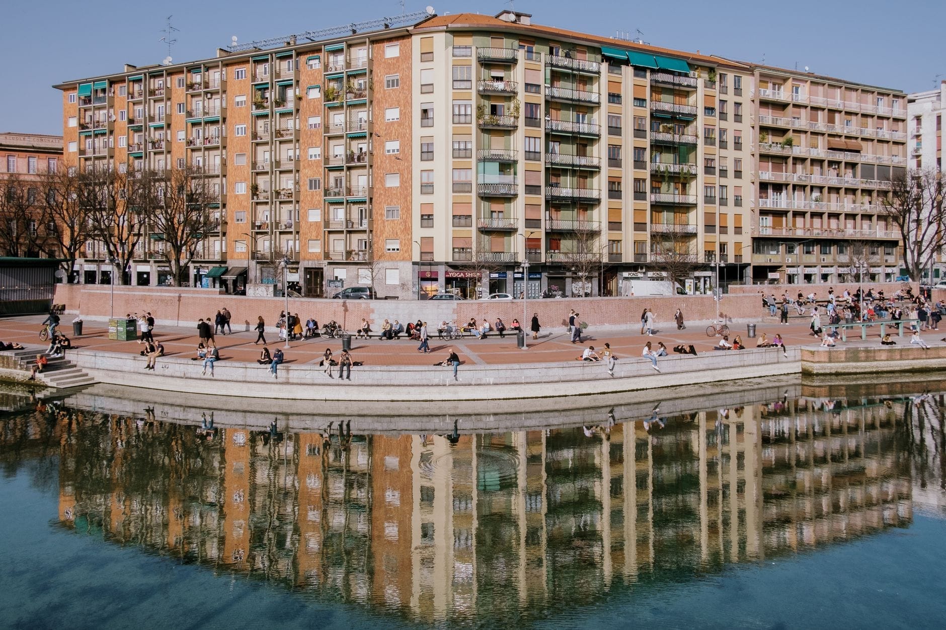 Le persone si rilassano sui gradini della Darsena di Milano, con colorati condomini e i loro riflessi visibili nel tranquillo bacino sul lungofiume.