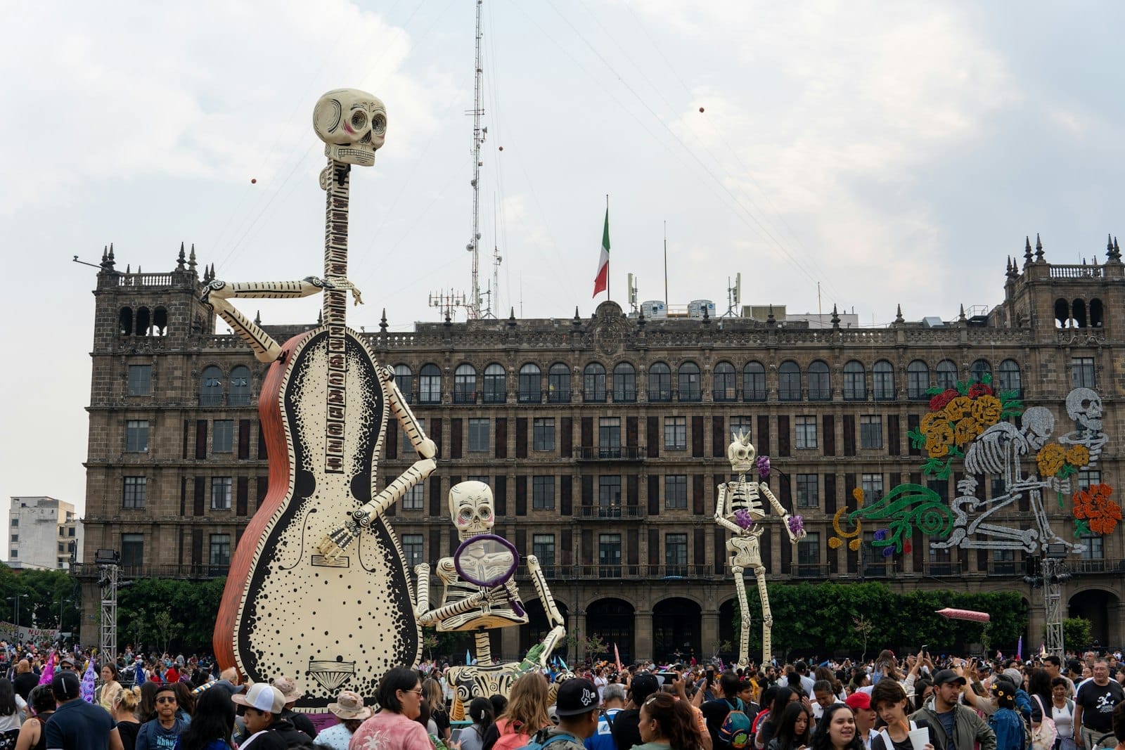 Zócalo lotado na Cidade do México com gigantes figuras de esqueletos do Día de Muertos e decorações em frente a um edifício histórico.