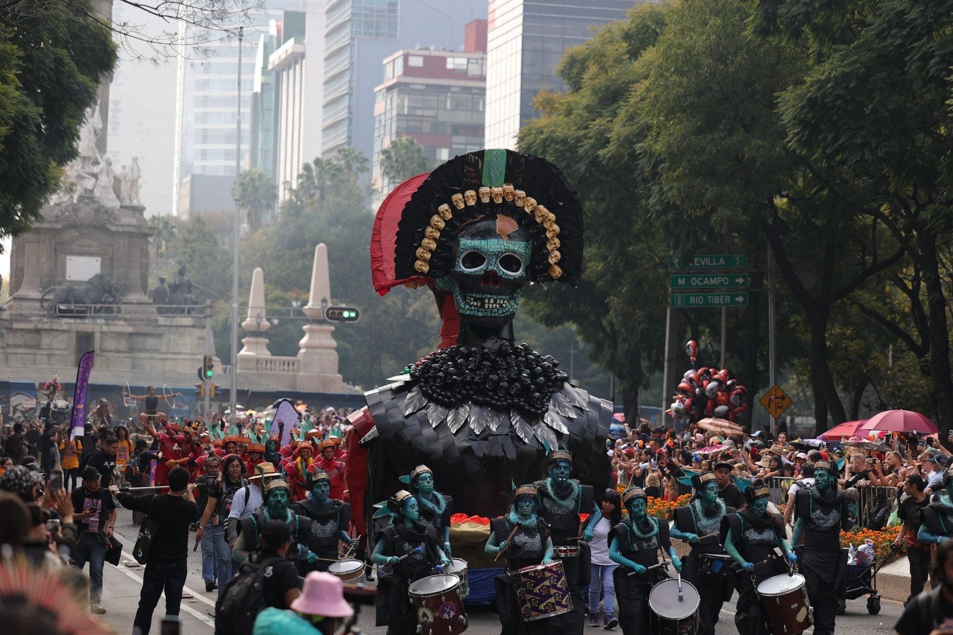 Desfile do Día de los Muertos na Cidade do México com um elaborado carro alegórico de esqueletos e grande multidão em uma avenida, cercada de edifícios e árvores.