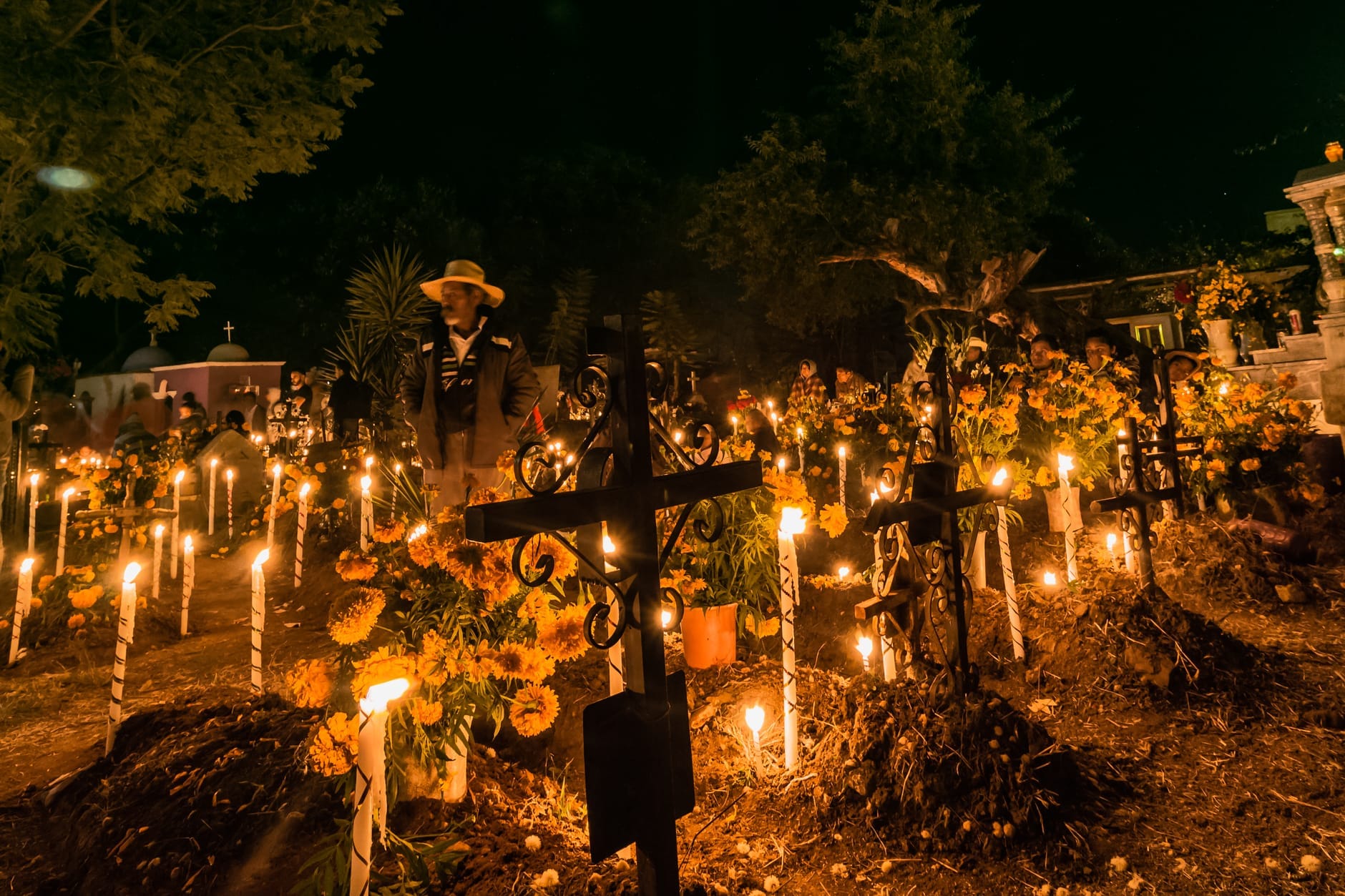 Cena noturna em um cemitério iluminado por velas com flores de calêndula, cruzes e pessoas observando as tradições do Día de los Muertos.