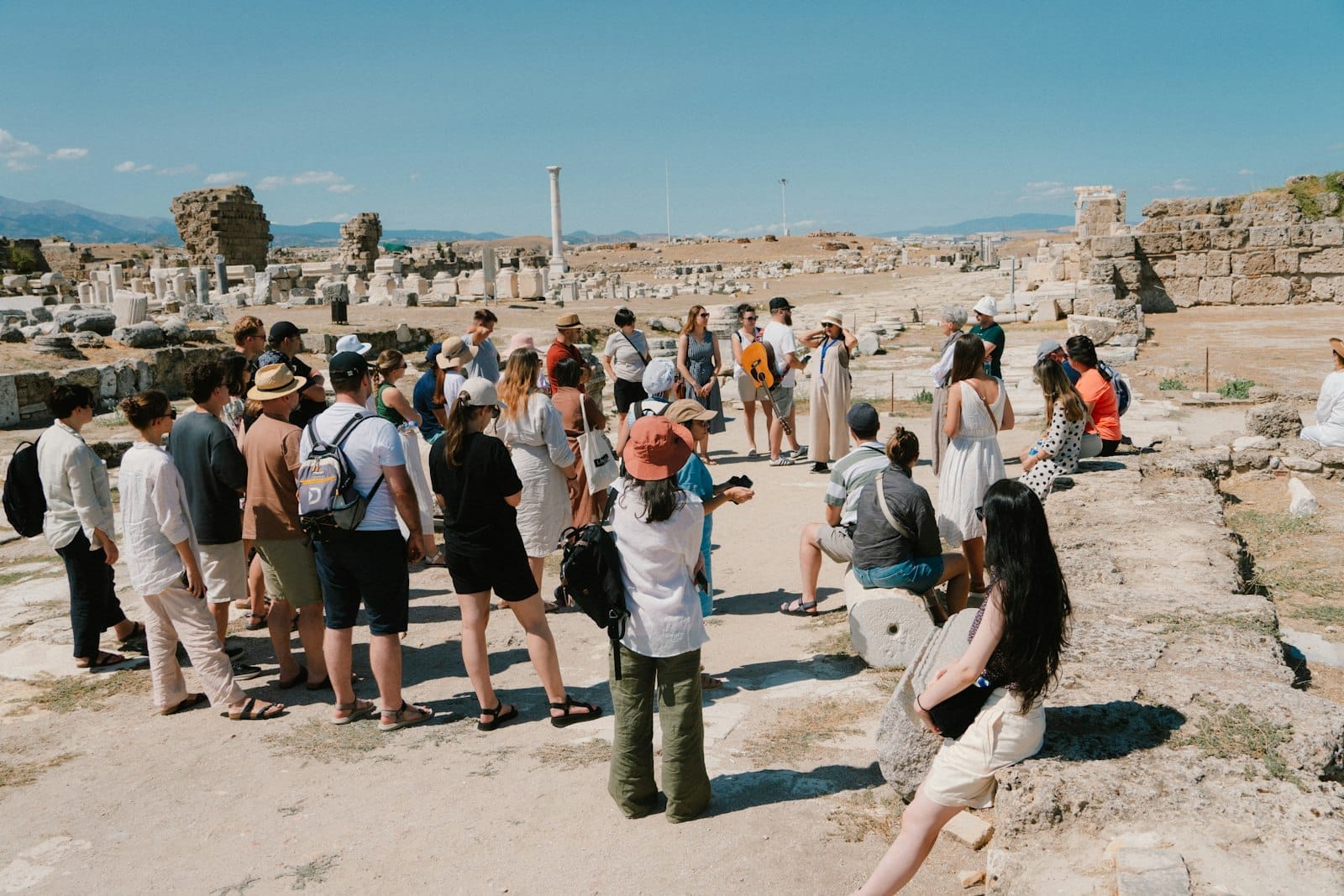 Un grupo de turistas se reúne alrededor de un guía entre ruinas antiguas y columnas de piedra bajo un cielo azul despejado