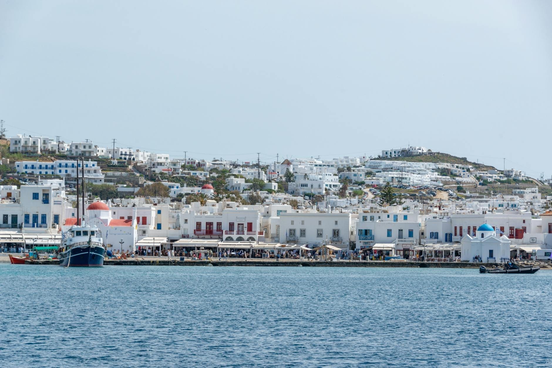 Vista del frente marítimo de Mykonos con barcos atracados, edificios encalados y la iglesia de cúpula azul de St. Nicholas cerca del punto de salida hacia Delos.