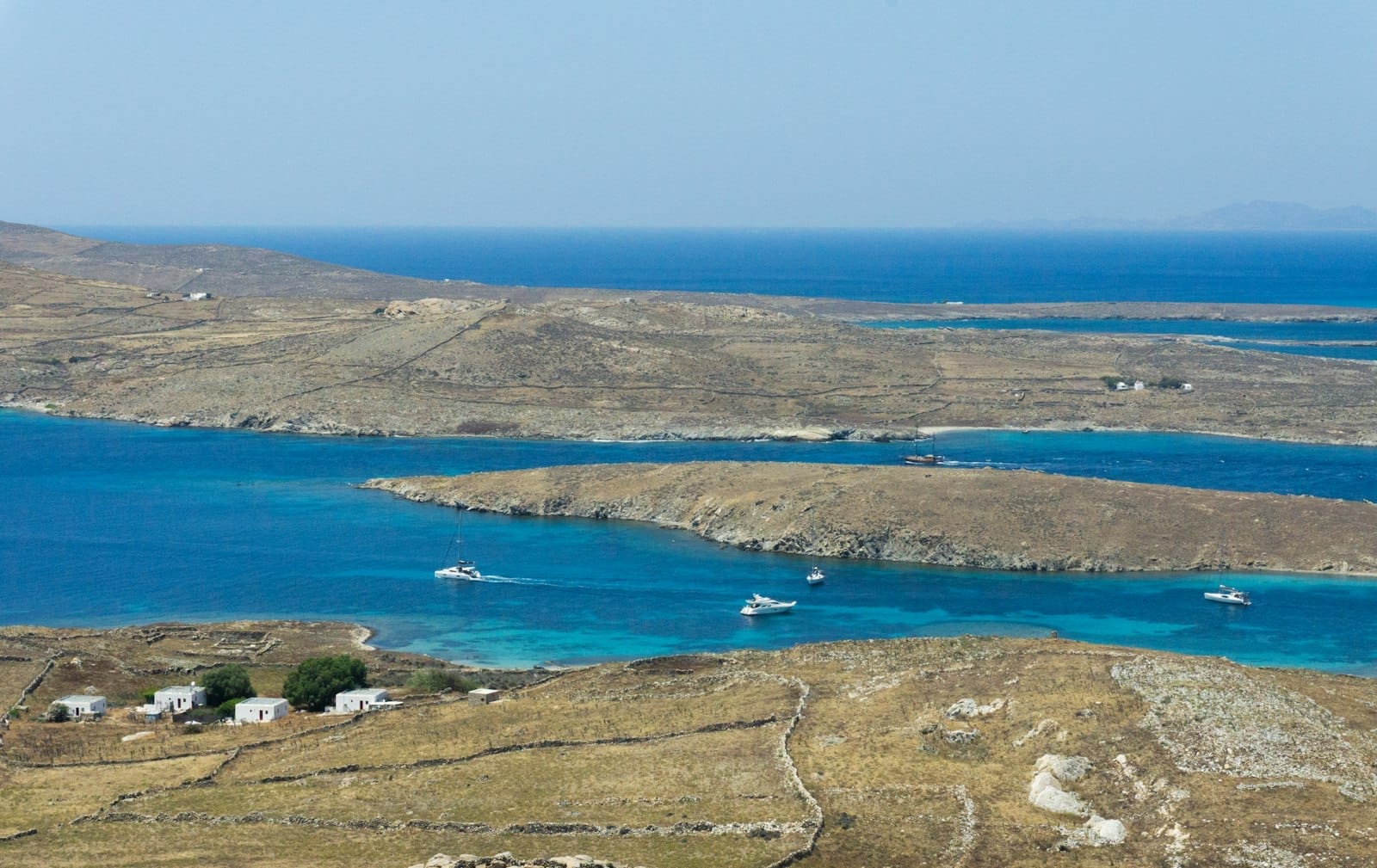 Vista panorámica de la árida isla de Delos rodeada por las profundas aguas azules del Egeo, con pequeñas embarcaciones navegando entre Delos y Mykonos bajo un cielo despejado.