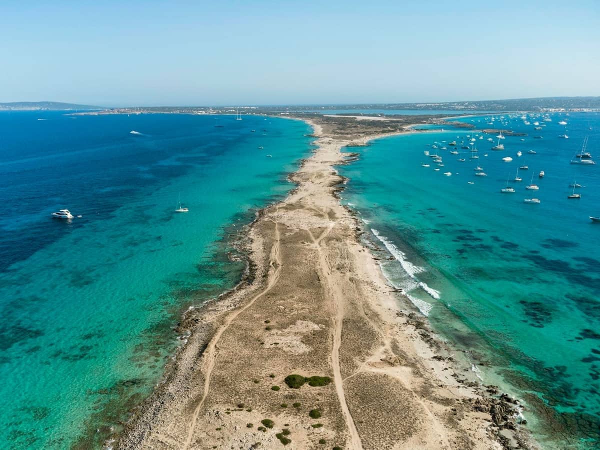 Aerial view of a narrow sandy strip surrounded by incredibly clear turquoise water and anchored boats, capturing Formentera’s pristine beach scene.