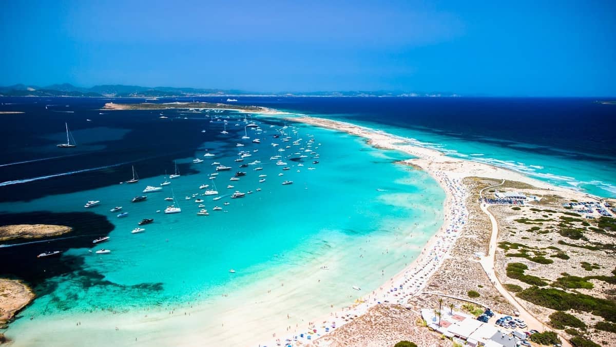 Aerial view of Ses Illetes beach in Formentera with turquoise waters, crowded sandy shore, and many anchored boats offshore on a sunny summer day.