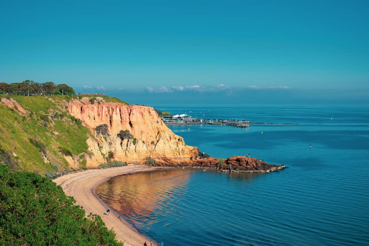 Golden cliffs, sandy beach, calm blue sea and a pier, typical of South Australia’s Fleurieu Peninsula coastline.