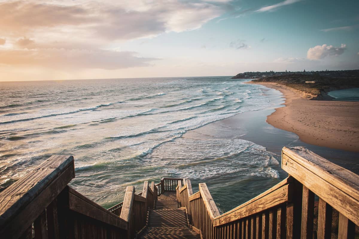 Wooden stairs leading down to a wide, sandy beach bordered by the blue waters and rugged coastline of the Fleurieu Peninsula near Adelaide.