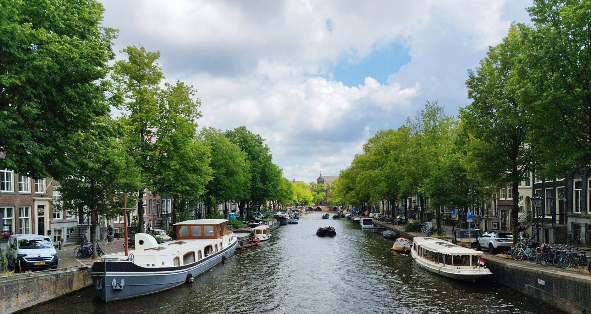 Vista amplia de un pintoresco canal de Ámsterdam bordeado de árboles y casas flotantes bajo un cielo parcialmente nublado. La escena invita a explorar.