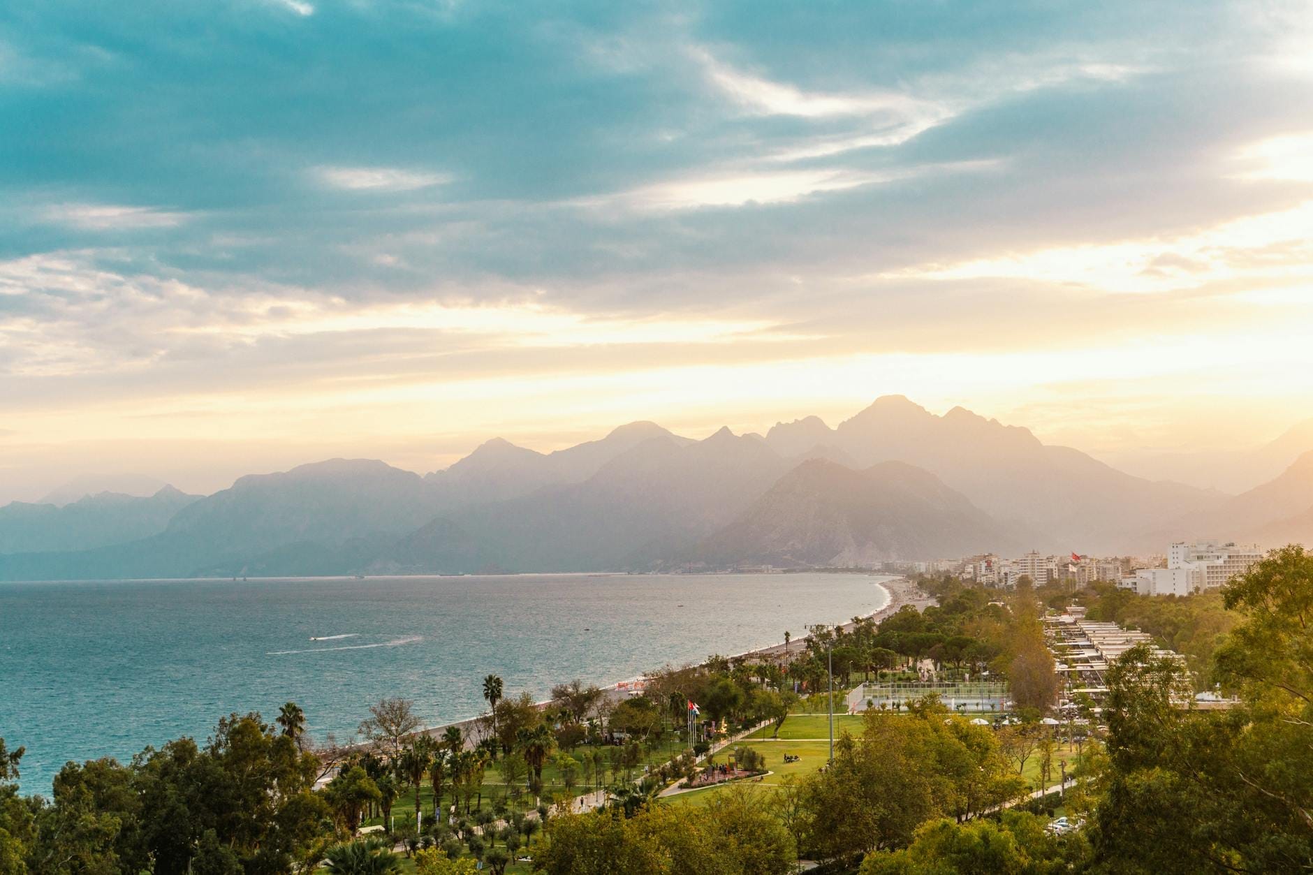 Vista panorâmica do litoral de Antalya ao pôr do sol, com montanhas imponentes, parques verdes e o Mar Mediterrâneo sob um céu colorido.