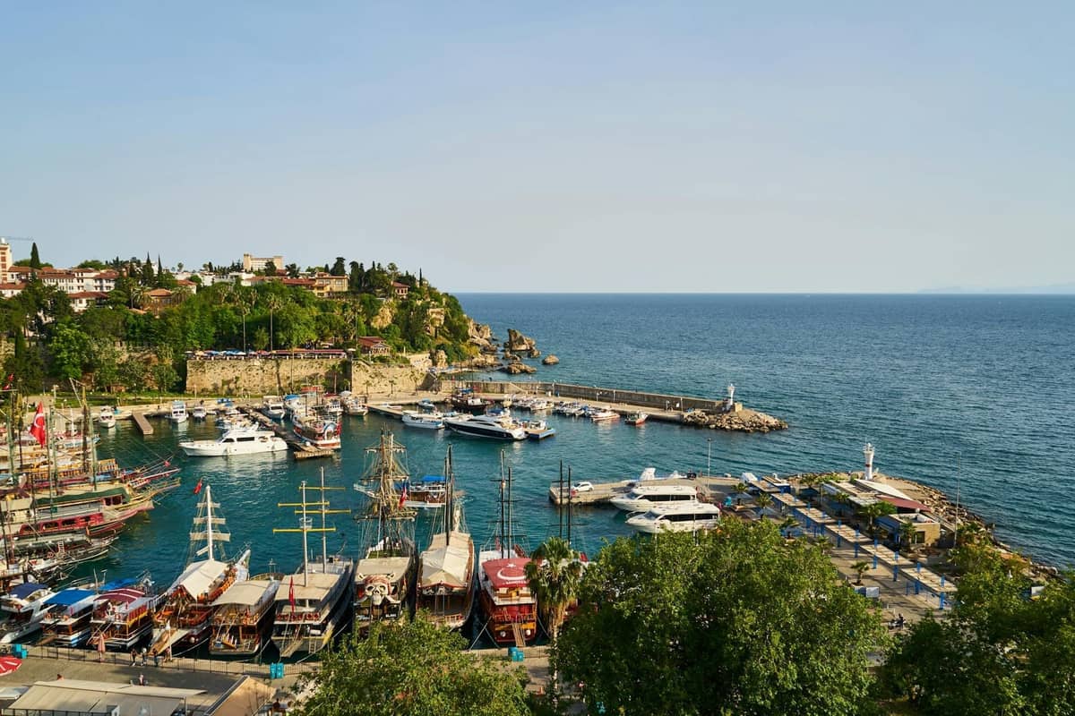 Scenic harbor in Antalya with boats, yachts, and historic old town buildings on a cliff beside the Mediterranean Sea under a clear sky.
