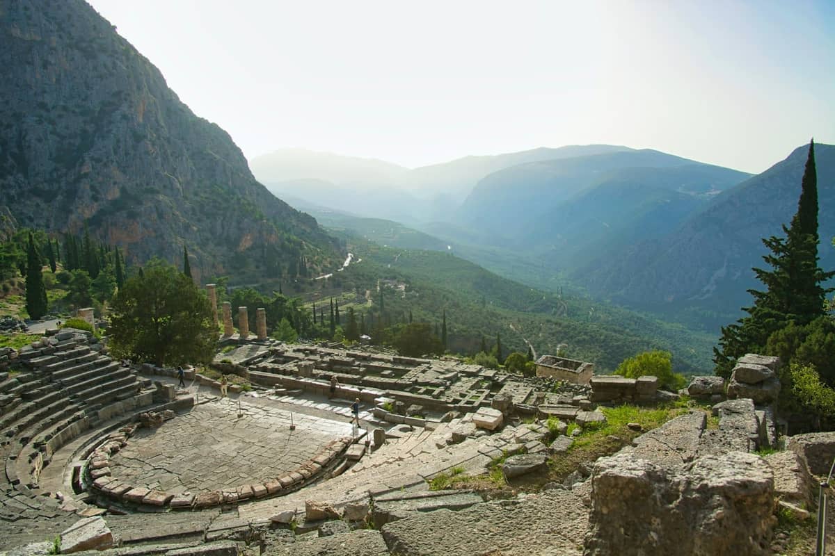 Panoramablick auf ein antikes griechisches Theater an einem Hang, umgeben von Bergen und Grün unter einem diesigen Himmel.