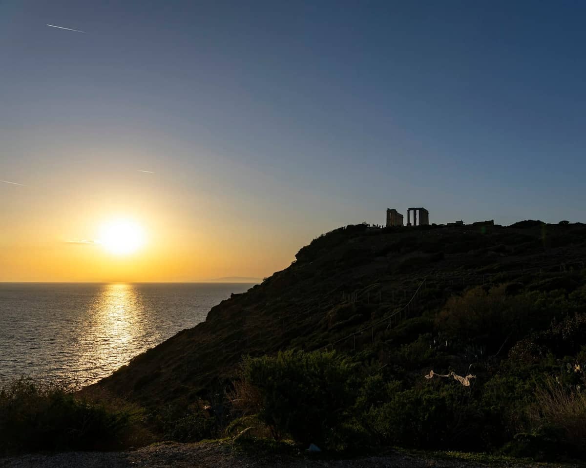 Sonnenuntergang entlang der Athenischen Riviera mit Cape Sounion und dem Tempel des Poseidon als Silhouette auf einem Küstenhügel über dem Meer.