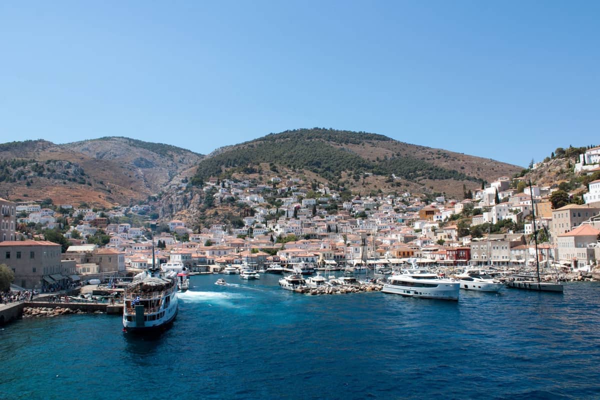 Eine lebhafte Hafenszene auf einer griechischen Insel mit weiß getünchten Häusern, im blauen Meer vertäuten Booten und Fähren sowie dahinter aufragenden Hügeln.
