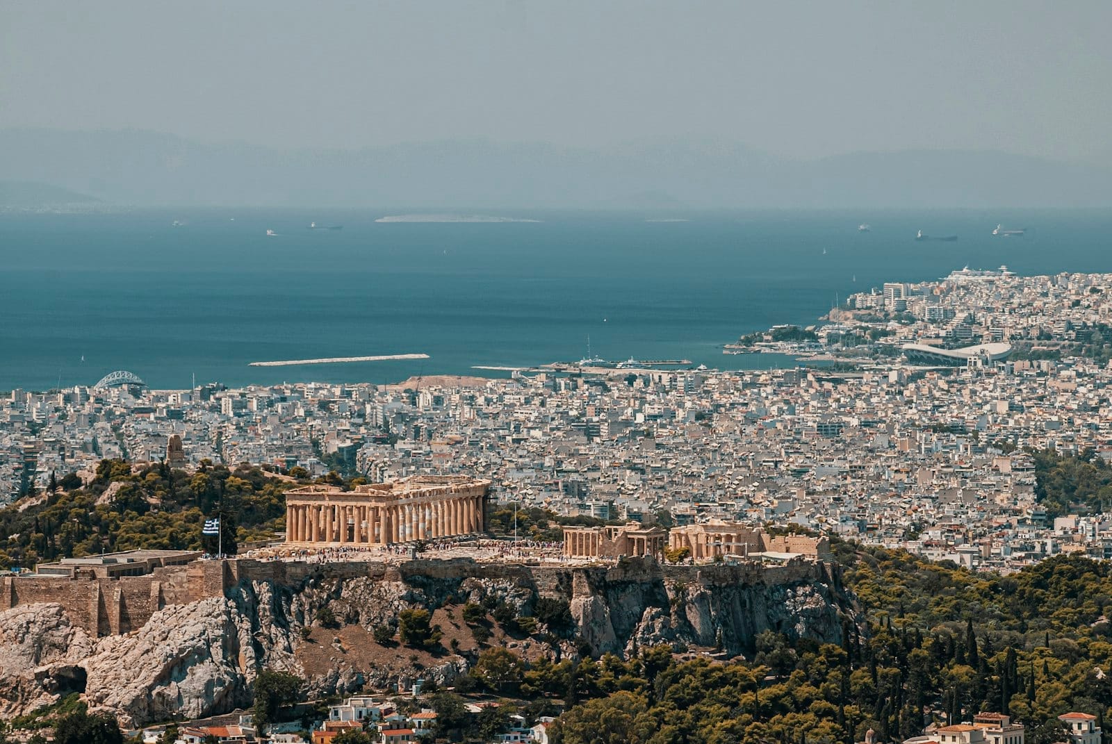 Vista aérea panorâmica de Atenas mostrando a Acrópole e o Partenon com a cidade se estendendo até o mar azul brilhante sob um céu limpo.