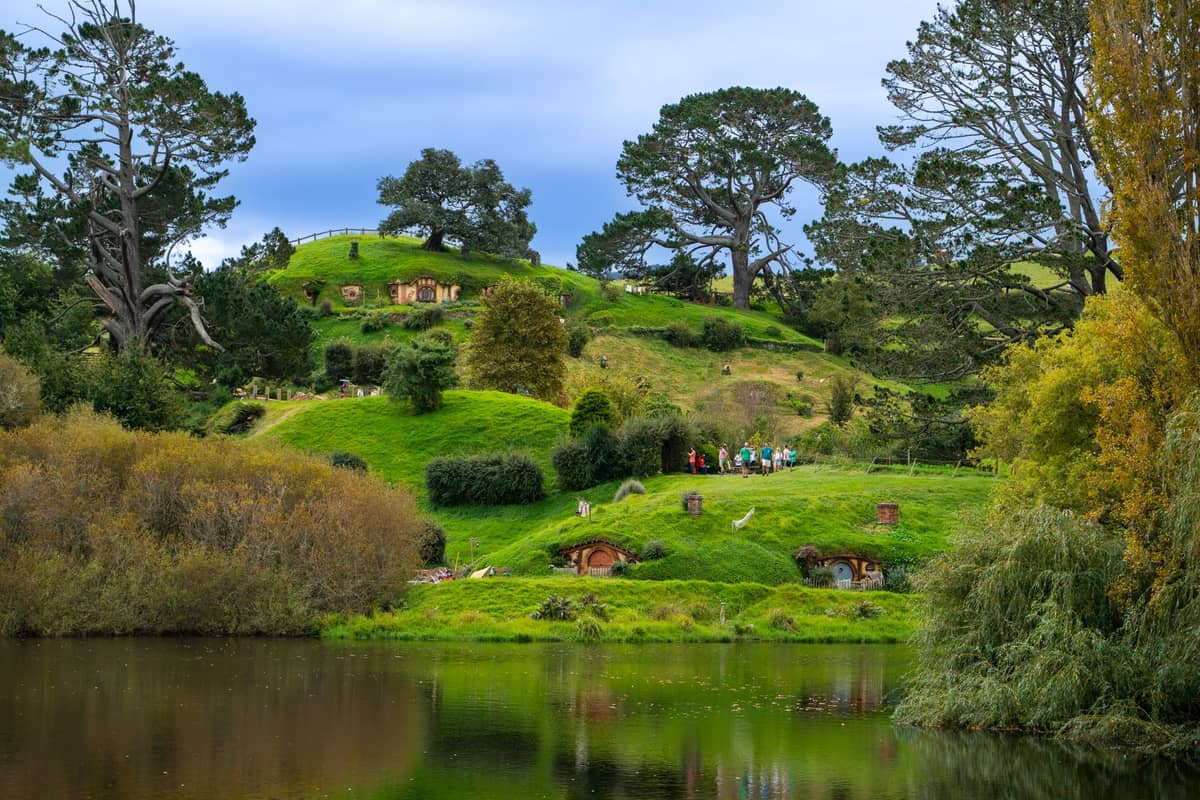 Szeroki widok na staw, trawiste wzgórza i okrągłe drzwi planu filmowego Hobbiton Movie Set, z grupą turystów widoczną wśród bujnego krajobrazu.