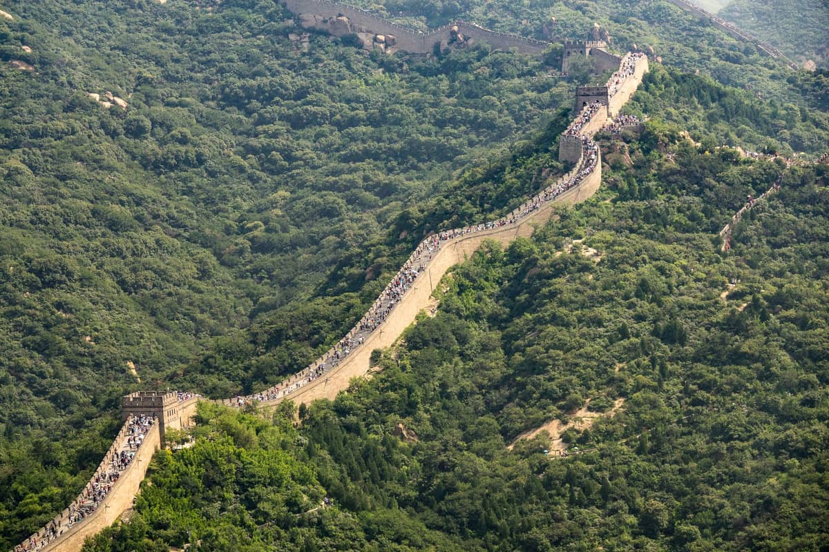 Vista aerea della Grande Muraglia Cinese che serpeggia su colline verdi affollate di visitatori, mettendo in risalto la lunghezza della muraglia e il suo paesaggio iconico.