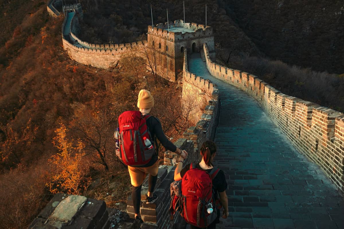 Zwei Wanderer mit Rucksäcken auf einem dramatischen Abschnitt der Chinesischen Mauer, umgeben von Bergen im goldenen Nachmittagslicht.
